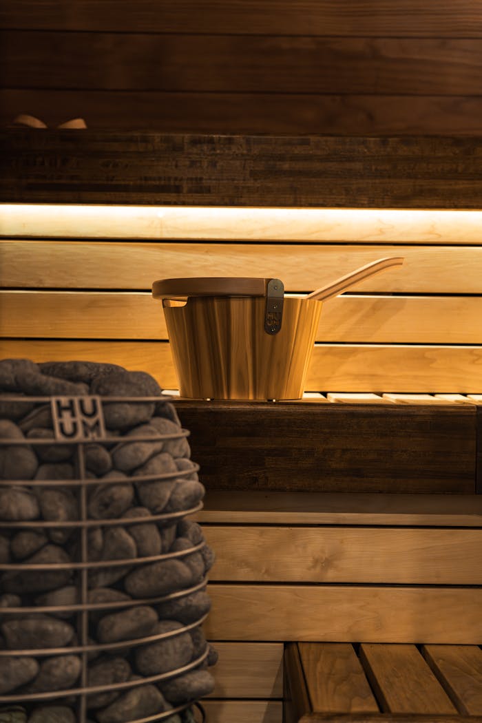 Warm lighting illuminates a Nordic wooden sauna featuring a bucket and heater stones, exuding relaxation.