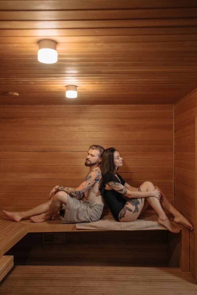 A tattooed couple relaxing back-to-back in a wooden sauna, enjoying a calming moment.